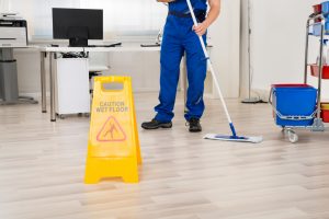 A person wearing a blue uniform using cleaning supplies to clean a floor. There's a "caution wet floor" sign up.