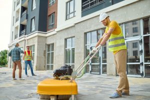 A man in high-vis work gear operating an industrial tile cleaning machine outdoors. In the background, two more men are looking at the building.
